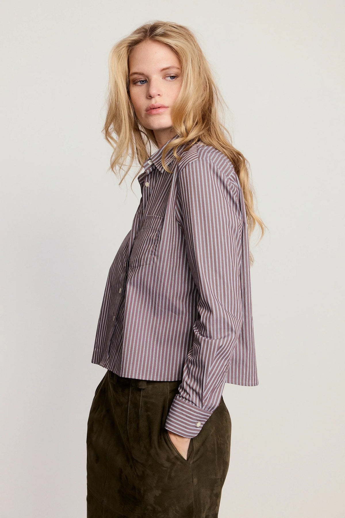 A woman wearing a long-sleeve button-up shirt with vertical purple and white stripes, paired with dark brown corduroy trousers, photographed against a light gray studio background.
