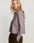 A woman wearing a long-sleeve button-up shirt with vertical purple and white stripes, paired with dark brown corduroy trousers, photographed against a light gray studio background.