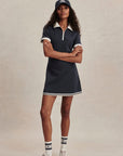 Woman wearing a navy dress with white trim, white sneakers, and a navy cap on a beige background