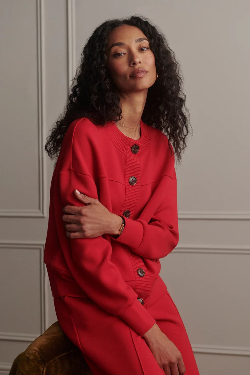 A woman with long, curly dark hair wearing a vibrant red knit cardigan dress with dark buttons, posed against a light gray paneled wall.