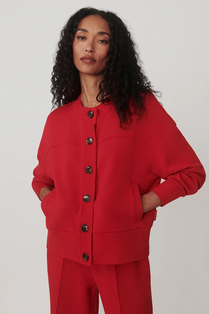 A model with curly black hair wearing a bright red cropped cardigan with dark buttons and matching red trousers, photographed against a neutral light gray studio background.