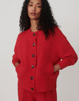 A model with curly black hair wearing a bright red cropped cardigan with dark buttons and matching red trousers, photographed against a neutral light gray studio background.