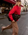 A woman walking on a city street wearing a bright red half-zip sweater, brown wide-leg trousers, a brown cap, and carrying a large black shoulder bag.