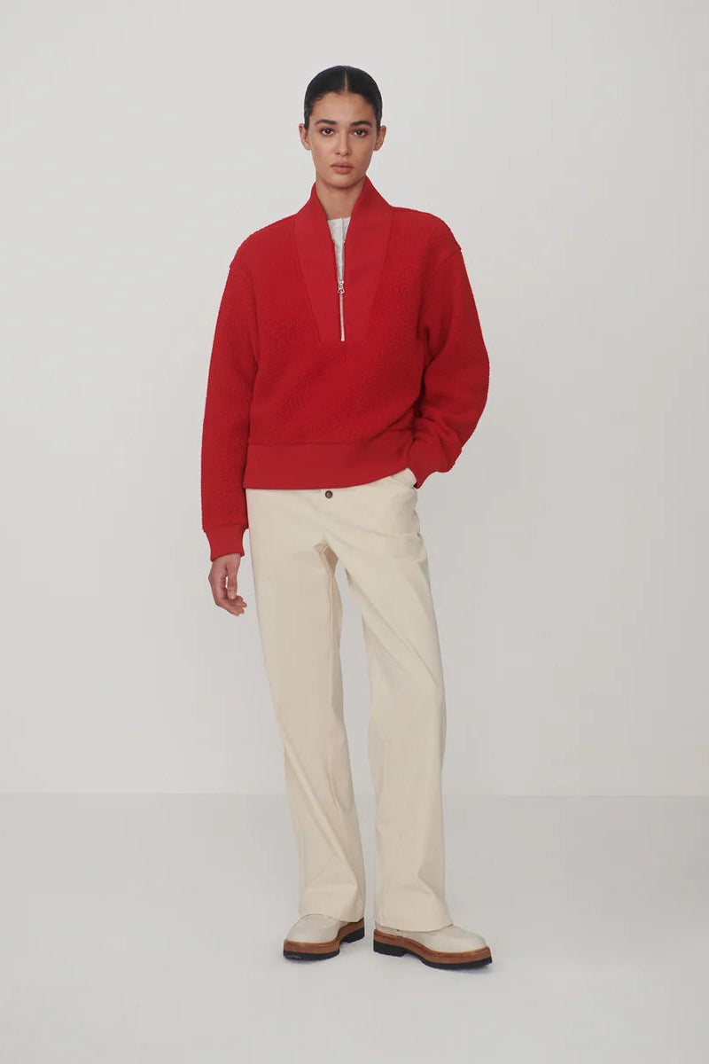A model stands against a light gray studio background wearing a bright red half-zip pullover sweater, cream-colored trousers, and brown-and-white shoes.