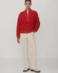 A model stands against a light gray studio background wearing a bright red half-zip pullover sweater, cream-colored trousers, and brown-and-white shoes.