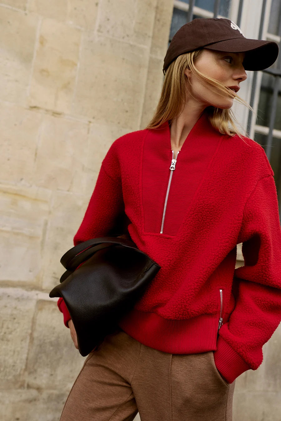 A woman wearing a bright red textured zip-up sweater, brown pants, a dark brown baseball cap, and holding a black leather bag, standing against a beige stone wall.