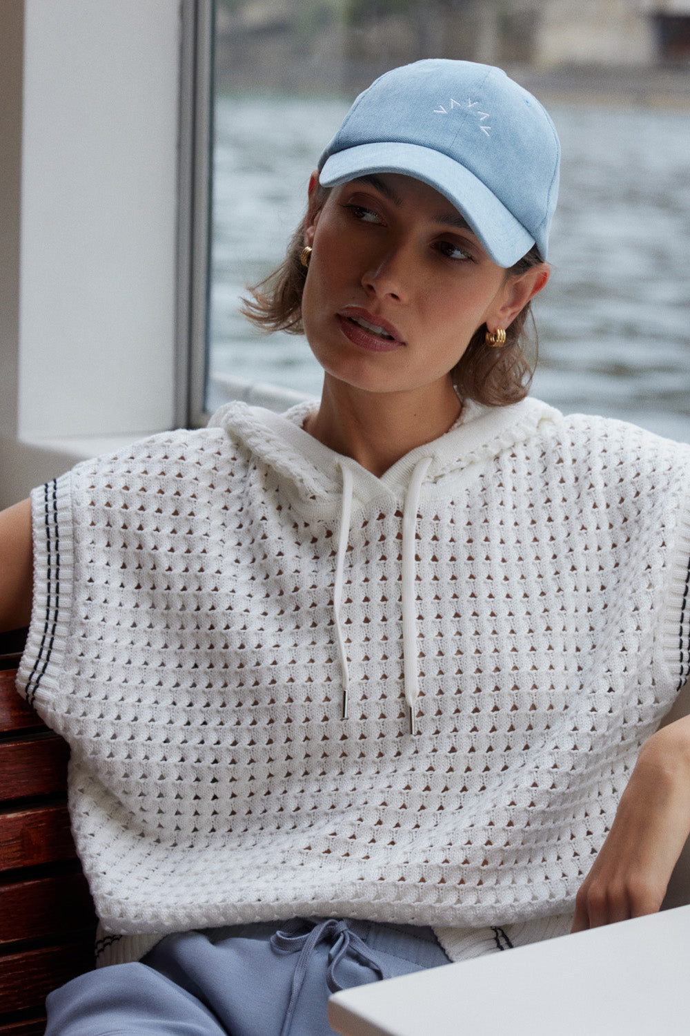 Woman wearing a light blue cap and sleeveless crochet top sitting by a waterfront.