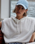 Woman wearing a light blue cap and sleeveless crochet top sitting by a waterfront.