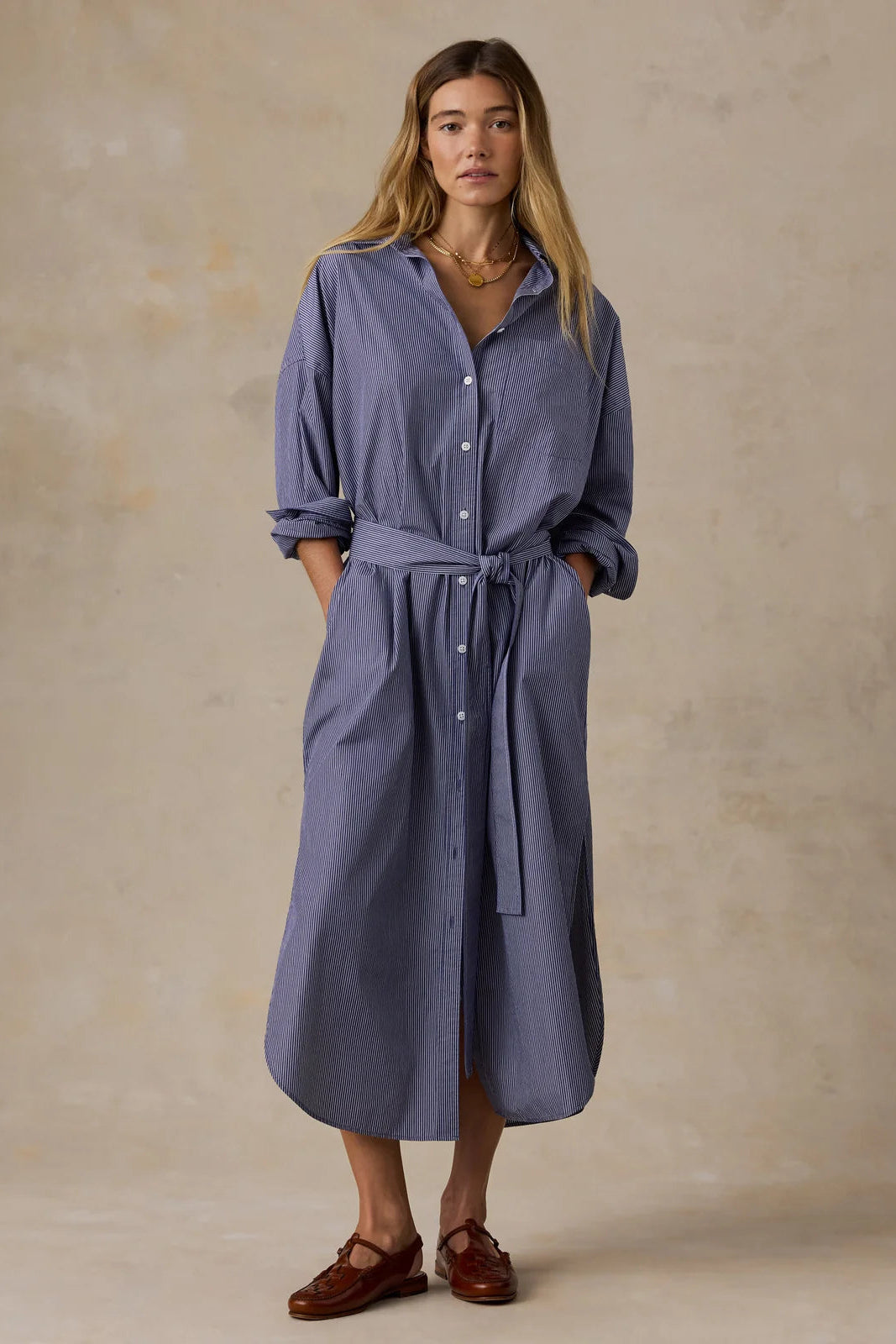A woman modeling a slate blue button-down shirt dress with a tie waist belt, standing against a textured beige studio backdrop.