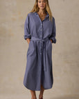 A woman modeling a slate blue button-down shirt dress with a tie waist belt, standing against a textured beige studio backdrop.