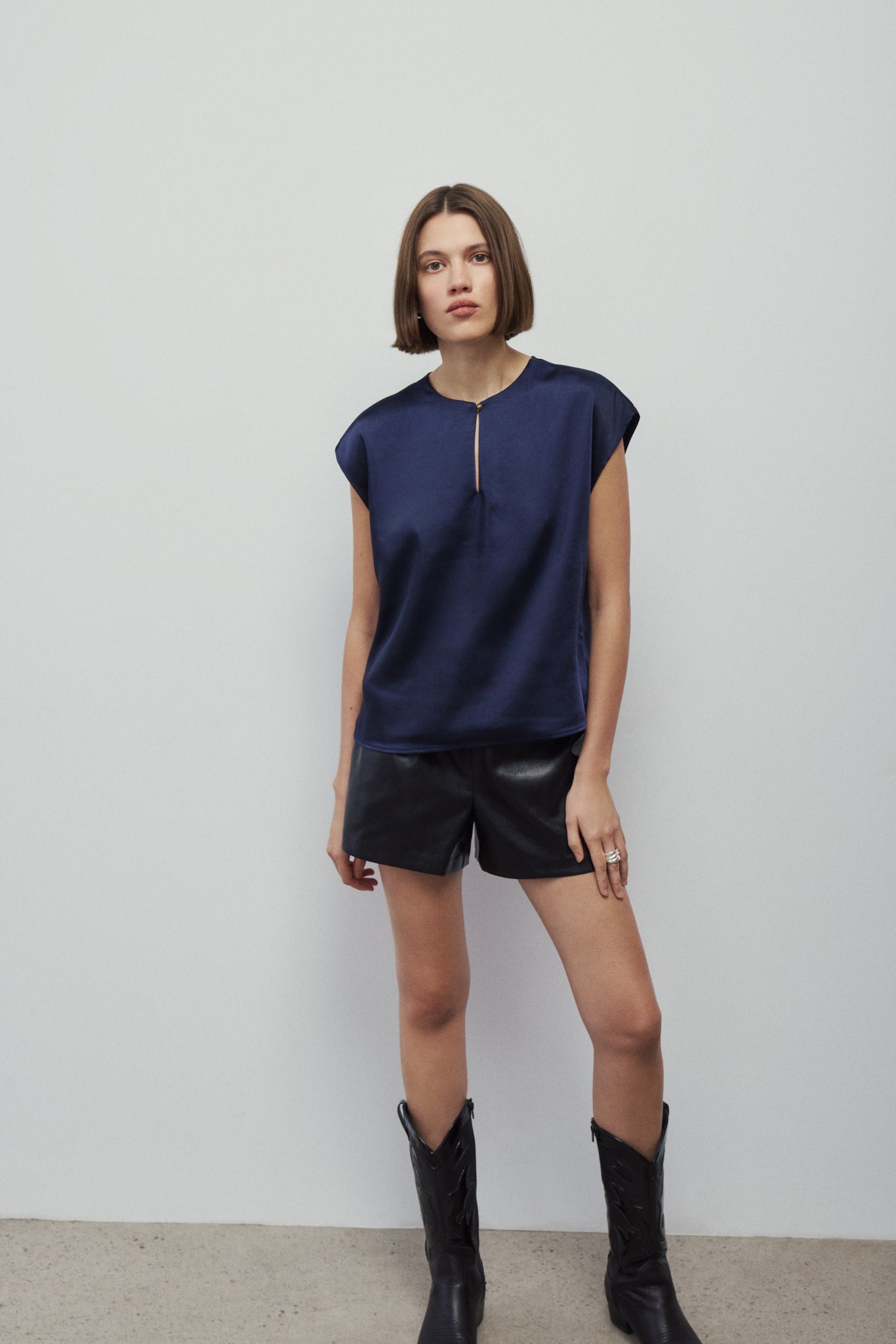 A woman modeling a navy blue short-sleeve top with a front zipper, paired with black shorts and black boots, standing against a plain light gray wall.