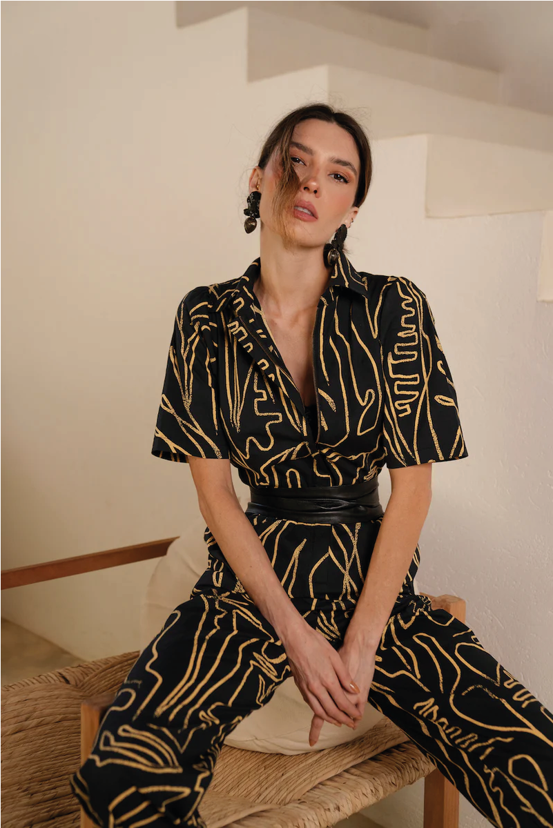 Woman wearing a black and gold patterned outfit sitting on a wooden stool.
