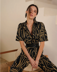 Woman wearing a black and gold patterned outfit sitting on a wooden stool.