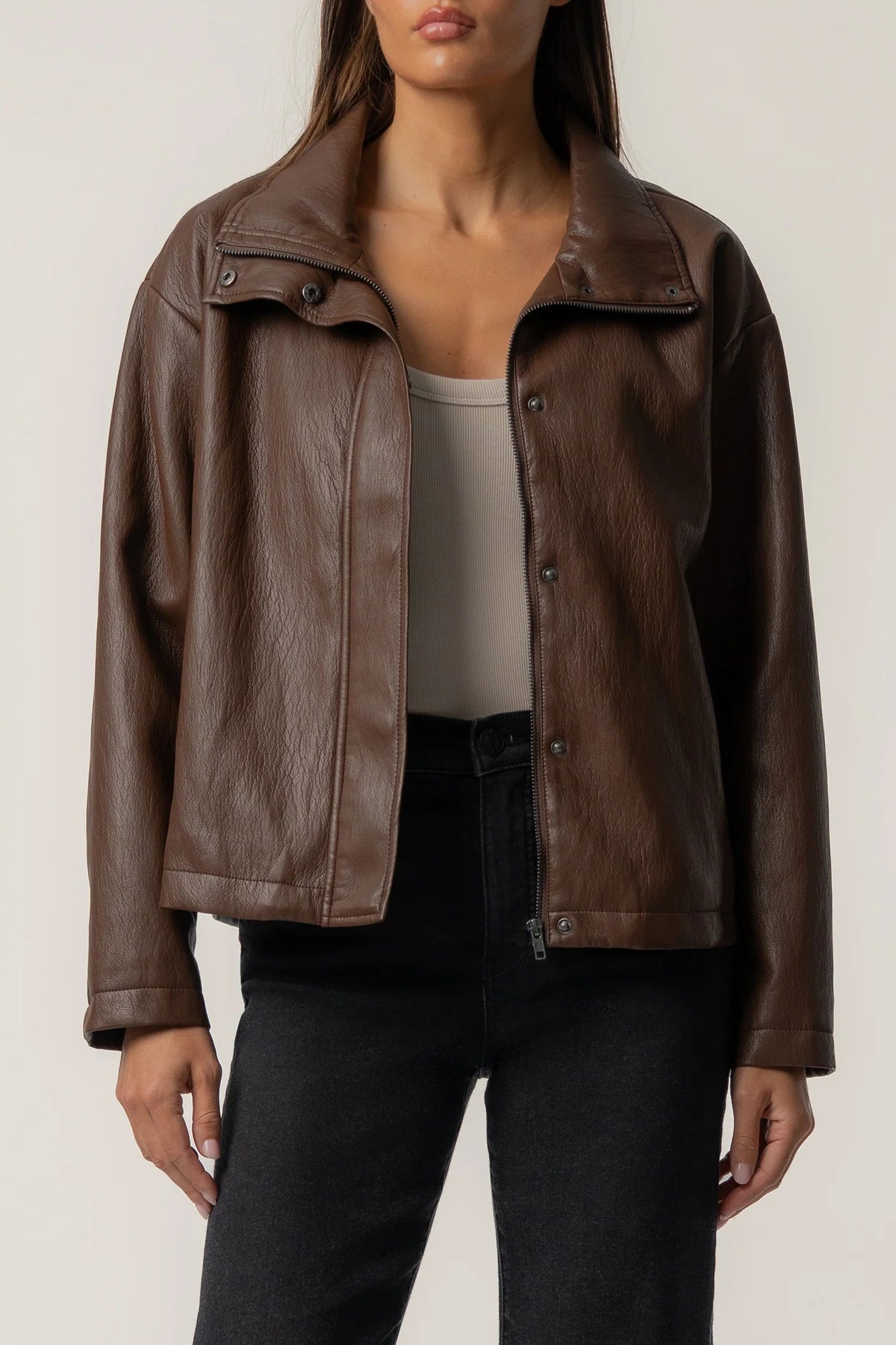 A woman wearing a brown faux-leather moto jacket with a relaxed fit, styled over a beige tank top and black jeans, against a plain light gray background.