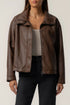 A woman wearing a brown faux-leather moto jacket with a relaxed fit, styled over a beige tank top and black jeans, against a plain light gray background.
