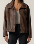 A woman wearing a brown faux-leather moto jacket with a relaxed fit, styled over a beige tank top and black jeans, against a plain light gray background.