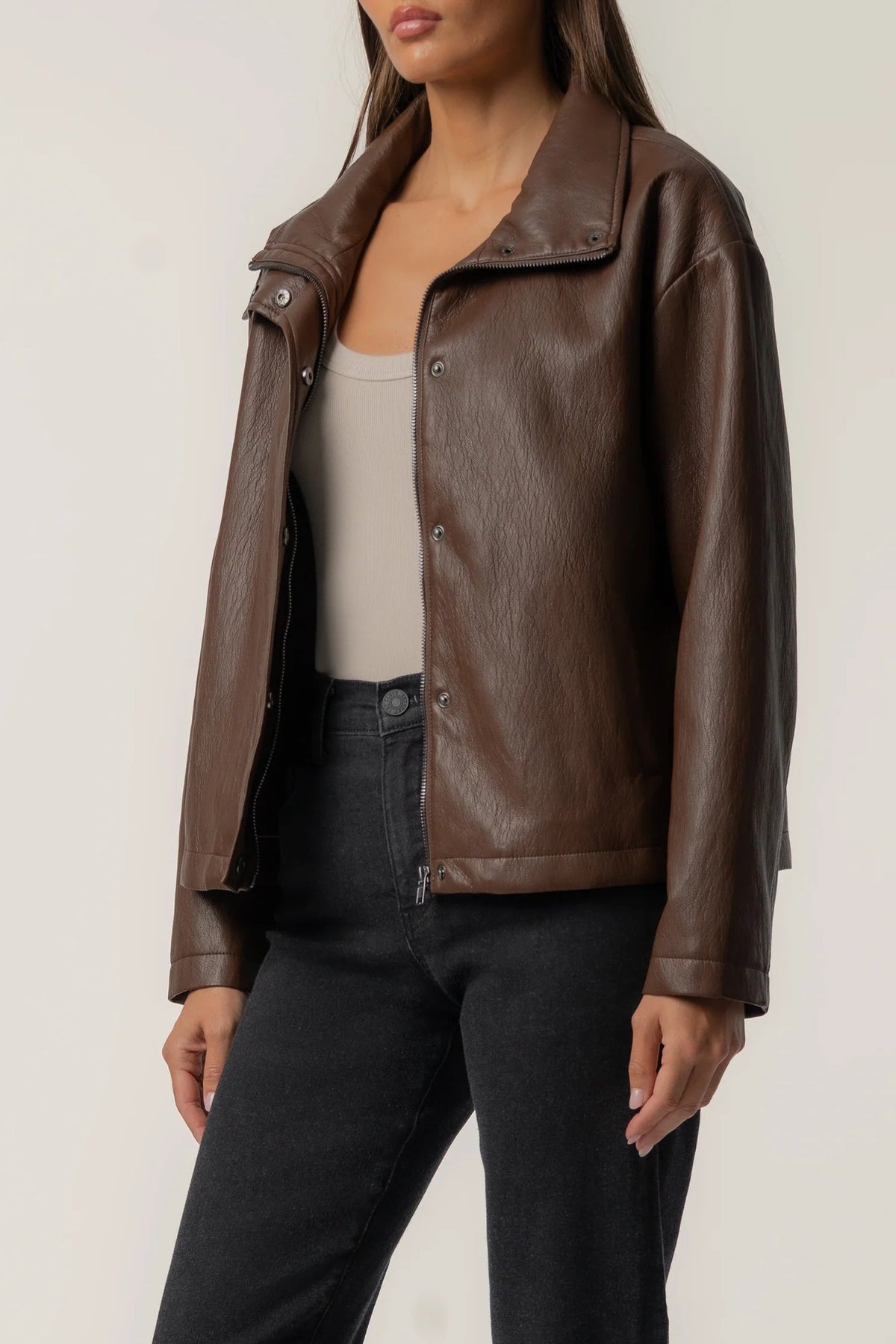 A woman wearing a brown faux-leather moto jacket over a beige tank top and black jeans, shown from the neck down against a plain light background.