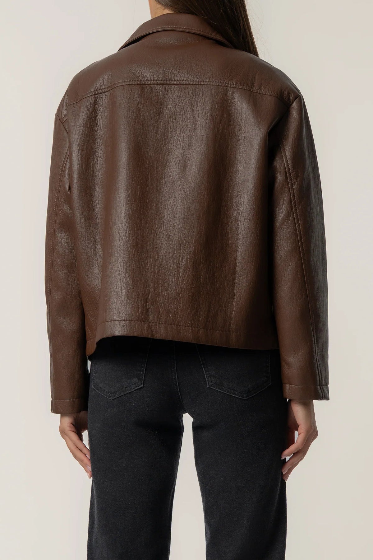 A person shown from the back wearing a cropped brown leather jacket and dark jeans against a plain light background.