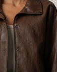 A close-up of a person wearing a dark brown faux-leather jacket with a collar, snap-button front, and zipper detail, paired with a light gray top against a plain light background.