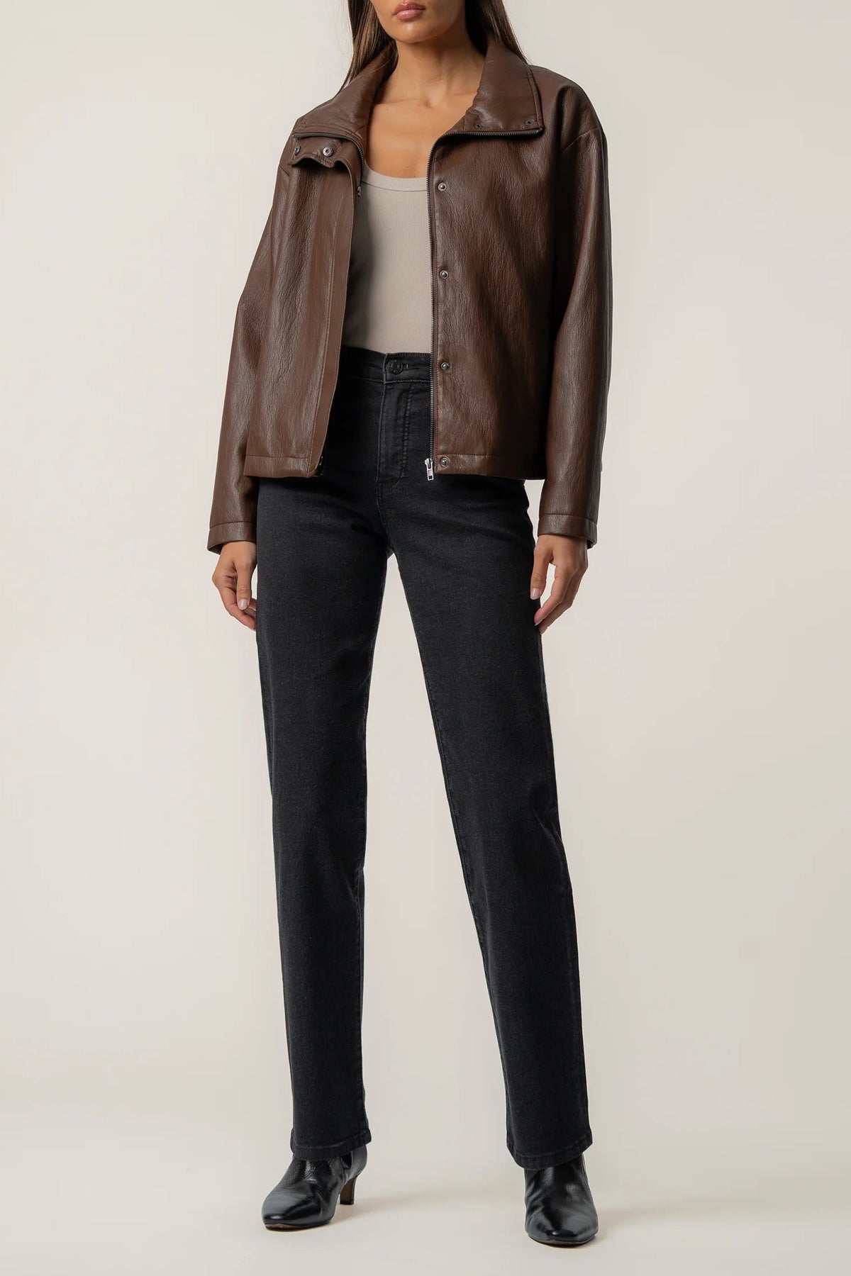 A model wearing a brown leather jacket, black jeans, and black ankle boots, standing against a plain light beige studio background.