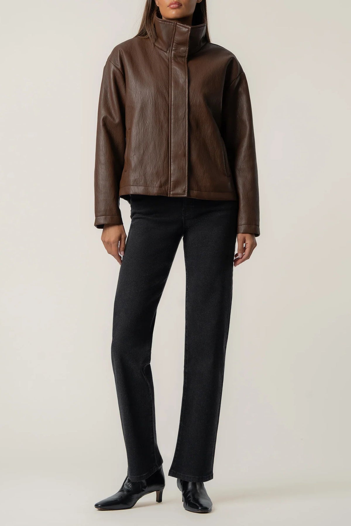 A model wearing a brown leather jacket with a high collar, paired with black trousers and black pointed-toe ankle boots, standing against a light beige studio background.