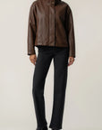 A model wearing a brown leather jacket with a high collar, paired with black trousers and black pointed-toe ankle boots, standing against a light beige studio background.