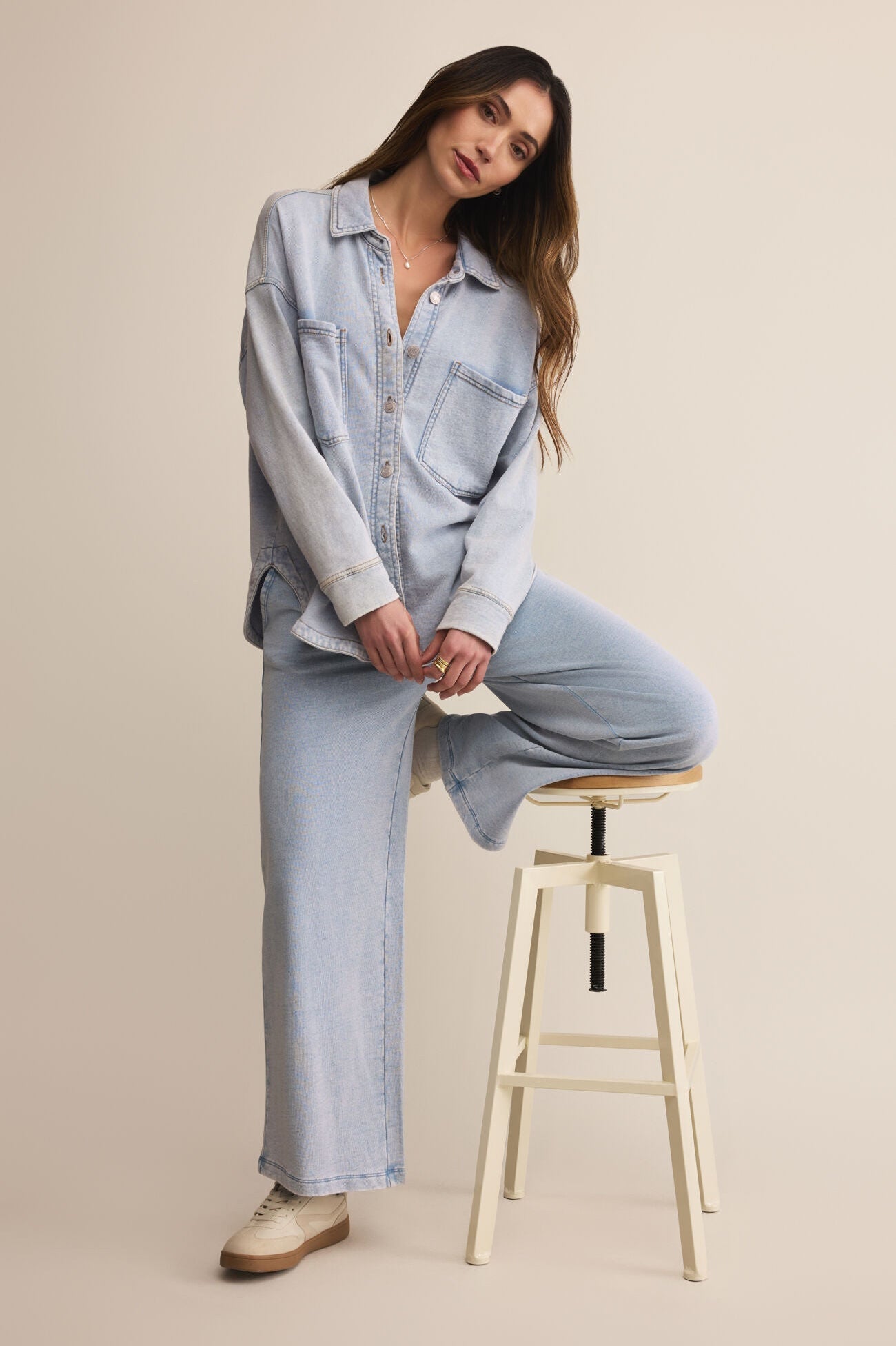 Woman sitting on a stool wearing a light blue denim outfit against a beige background