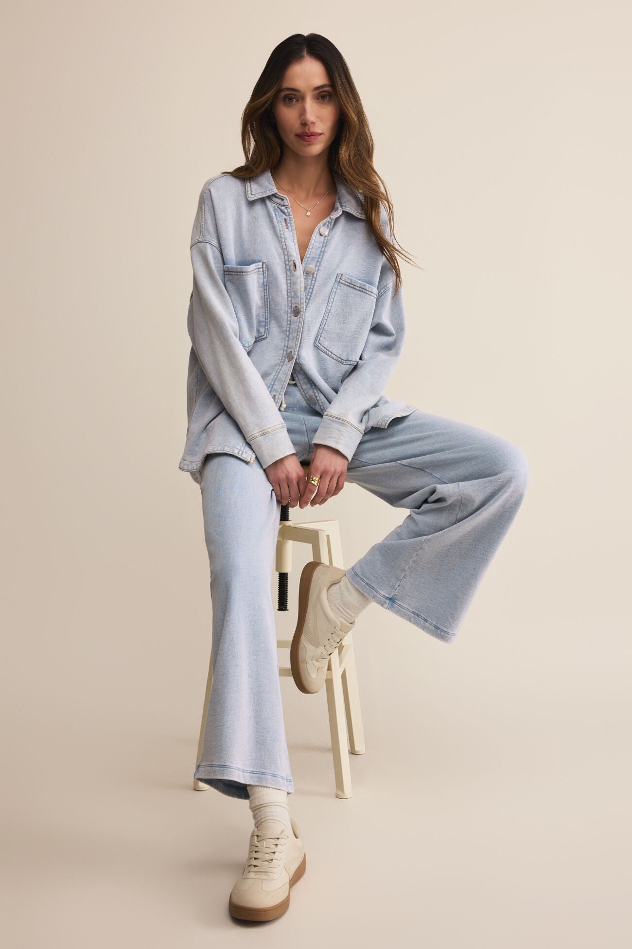 Woman sitting on a stool wearing a light blue denim outfit against a beige background