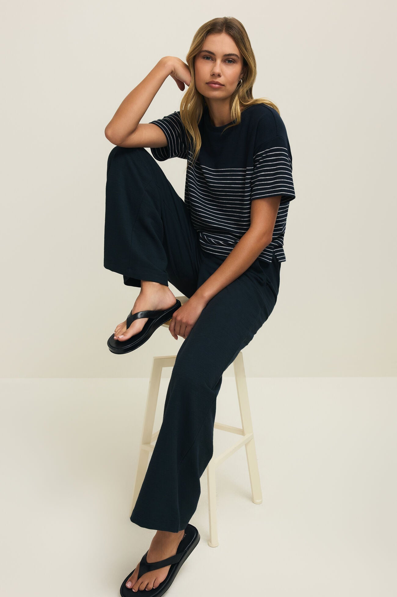 Woman sitting on a stool wearing a black and white striped shirt and dark pants against a plain background