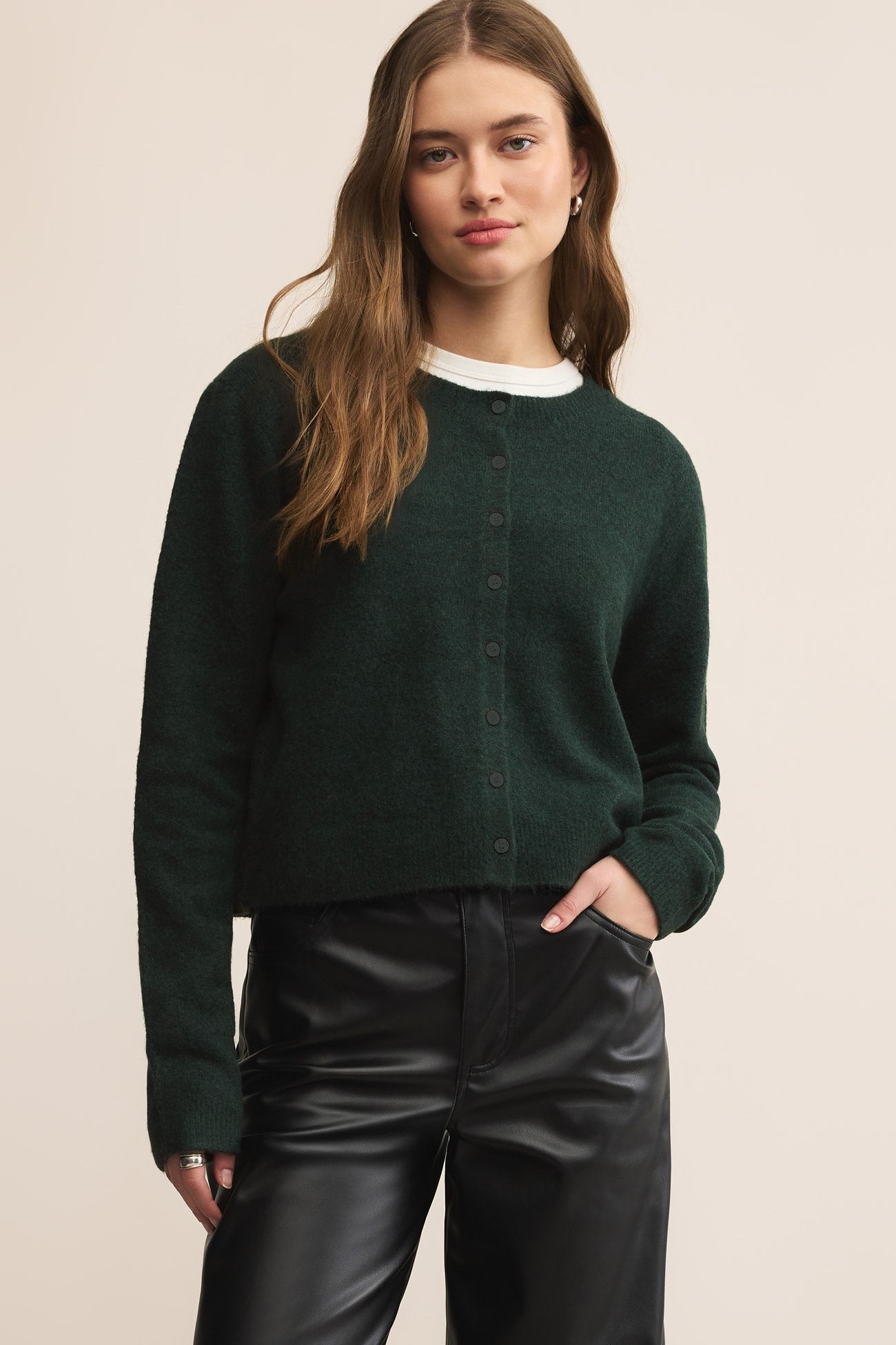Woman wearing a dark green cardigan over a white shirt and black pants on a beige background