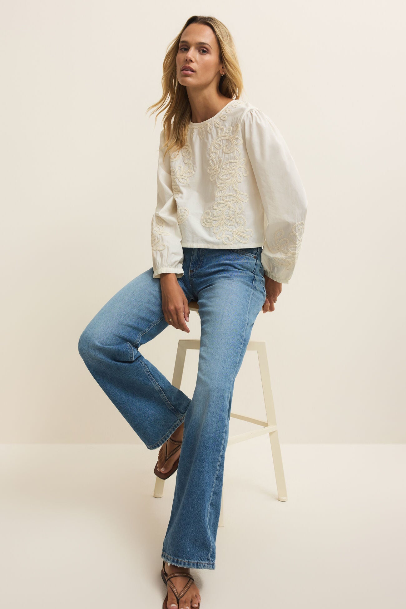 Woman wearing a white sweater and blue jeans sitting on a white stool against a plain background