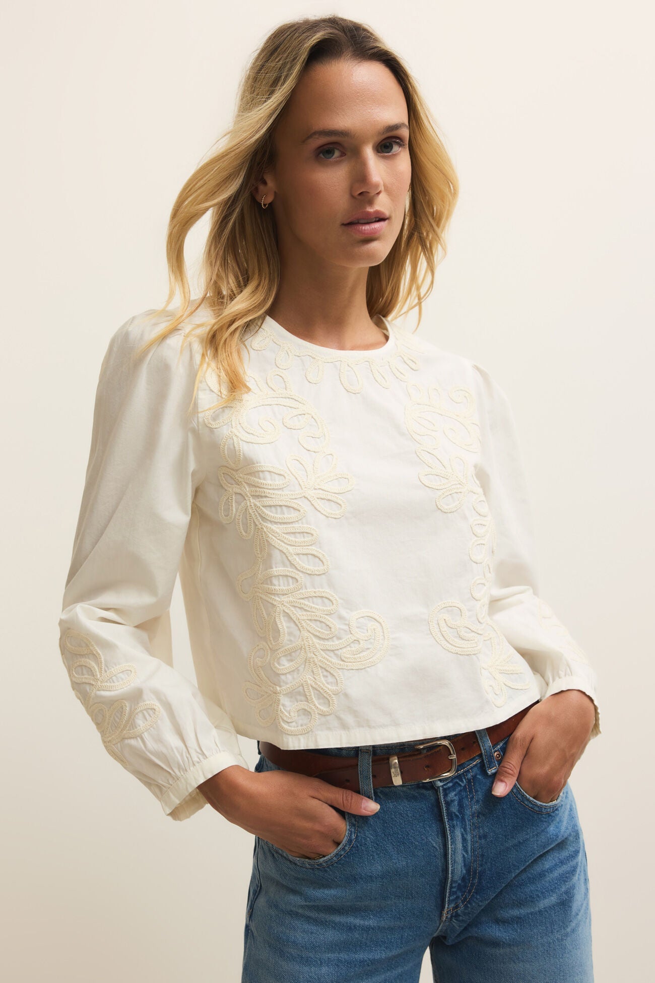 Woman wearing a white blouse with lace details and blue jeans against a plain background