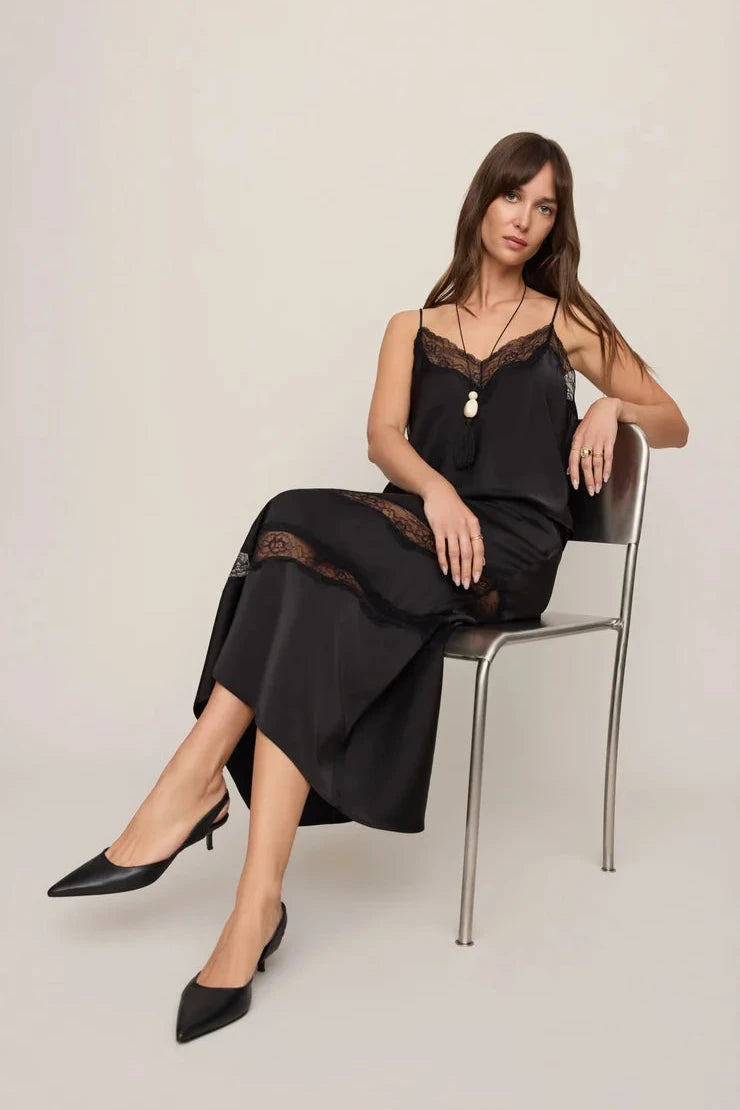 A woman sits on a metal chair wearing a black satin slip dress with lace trim and black pointed-toe heels against a plain light beige studio background.