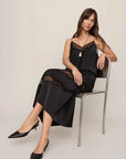 A woman sits on a metal chair wearing a black satin slip dress with lace trim and black pointed-toe heels against a plain light beige studio background.