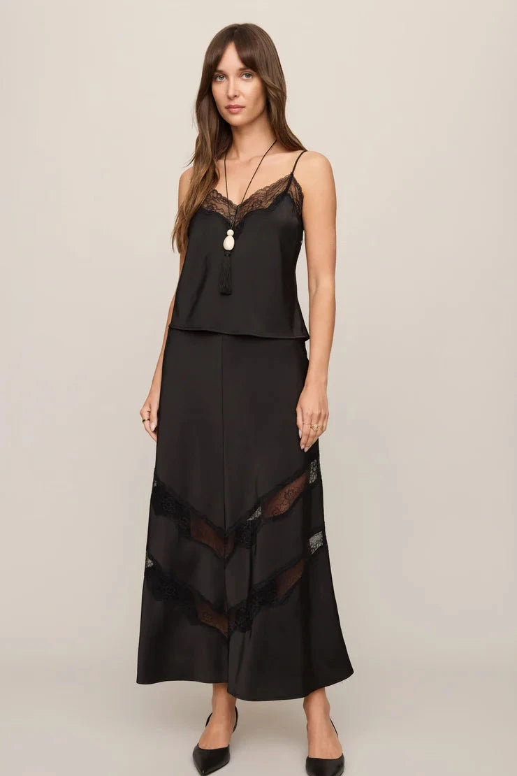 A woman modeling a black two-piece camisole and maxi skirt set with sheer lace panels, photographed against a plain light gray studio background.