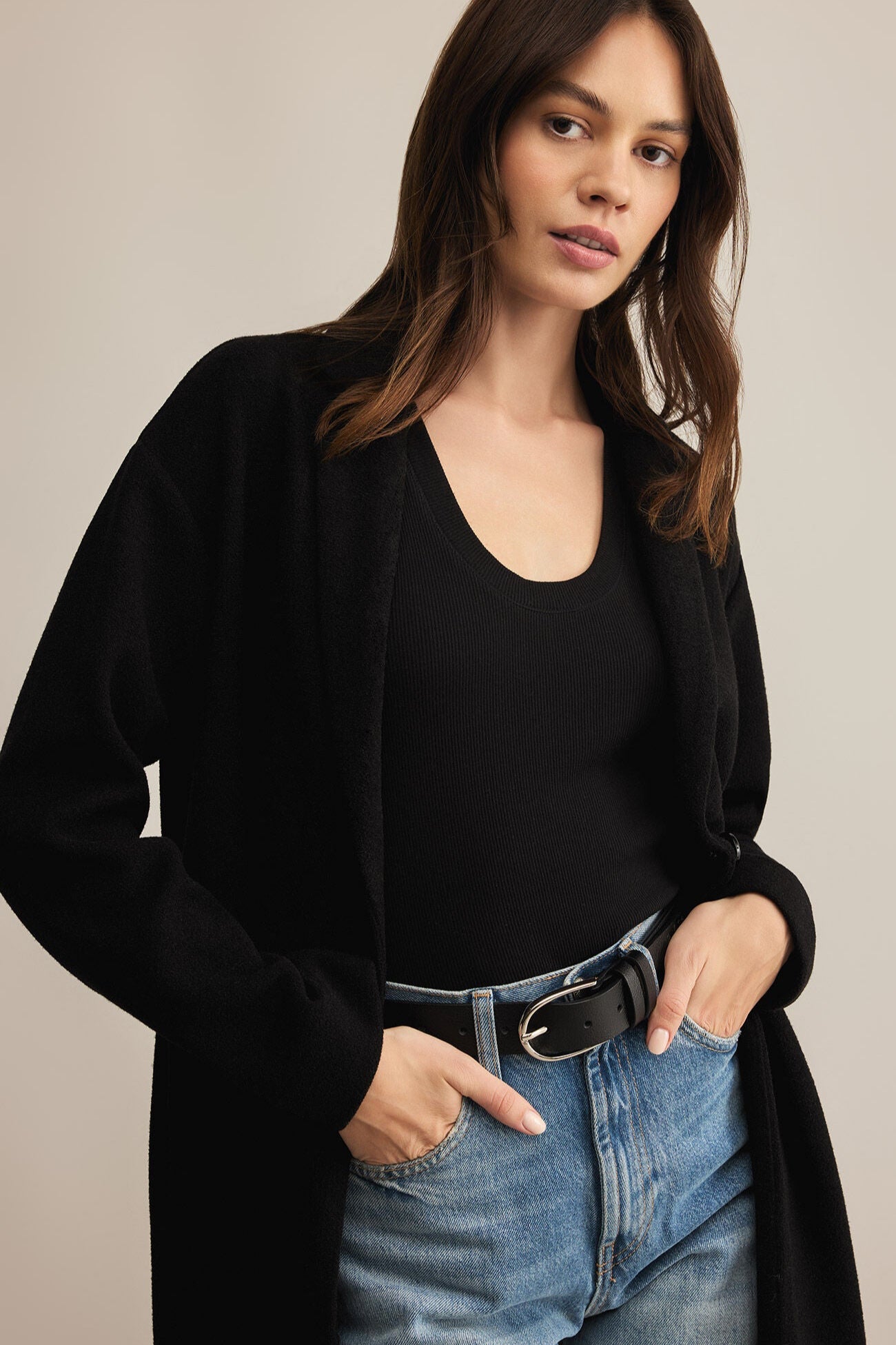 Woman wearing a black cardigan over a black top and blue jeans on a beige background