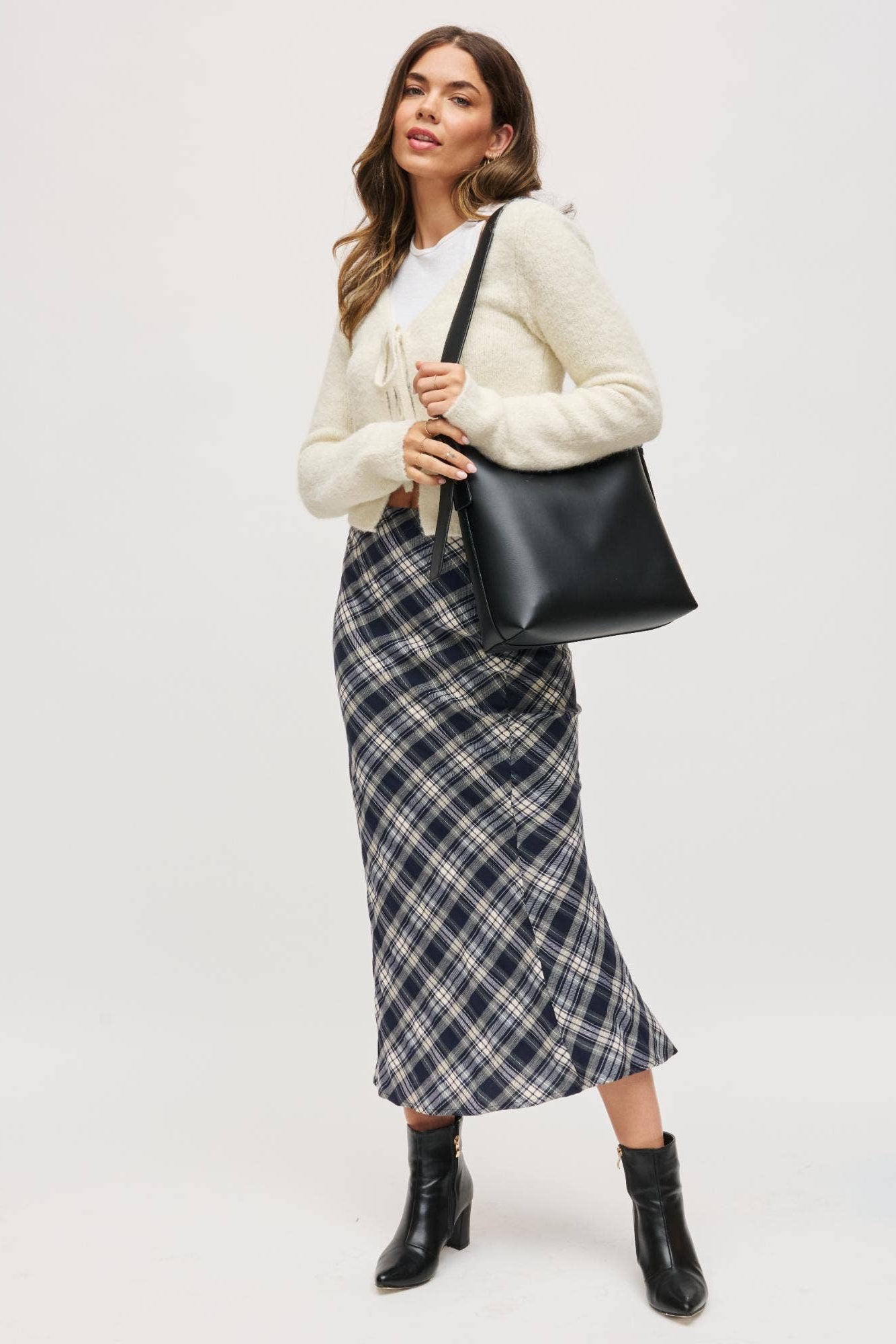 Woman holding a black handbag wearing a plaid skirt and white sweater on a white background