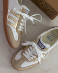 Pair of gold and white sneakers on a light surface with a box in the background.