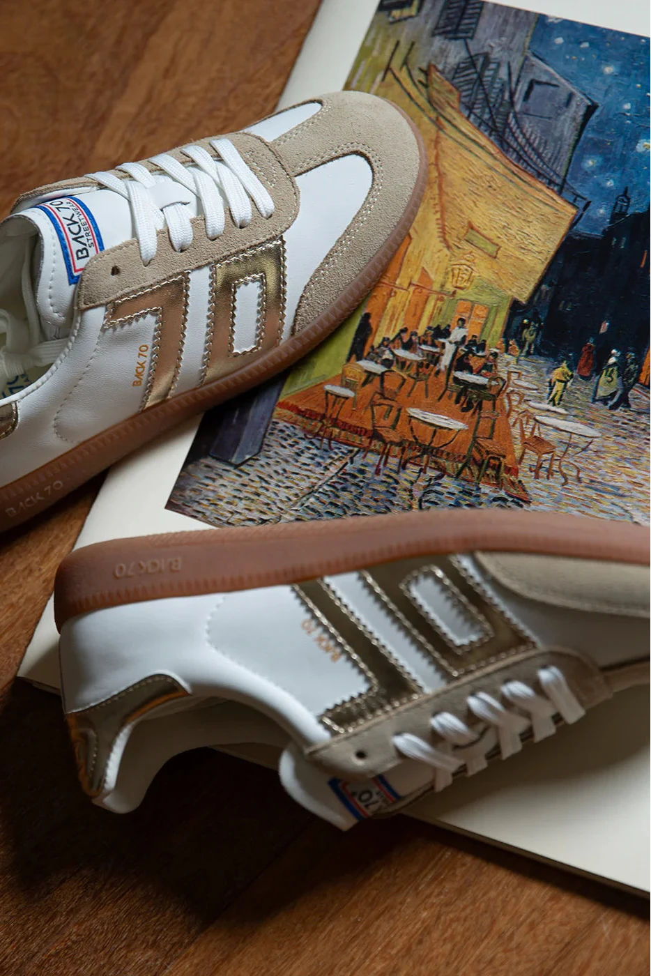 Pair of white sneakers with brown accents on a wooden surface with a painting in the background.
