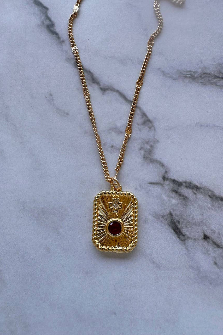 A gold chain necklace with a rectangular pendant featuring a sunburst design and a central dark red gemstone, displayed on a white marble surface.