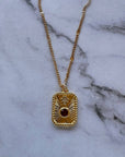 A gold chain necklace with a rectangular pendant featuring a sunburst design and a central dark red gemstone, displayed on a white marble surface.