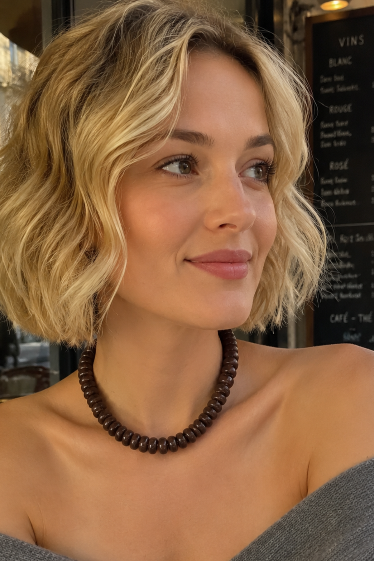 A woman with short blonde wavy hair wearing a dark brown beaded necklace and a gray off-the-shoulder sweater, sitting at a cafe.