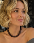 A woman with short blonde wavy hair wearing a dark brown beaded necklace and a gray off-the-shoulder sweater, sitting at a cafe.