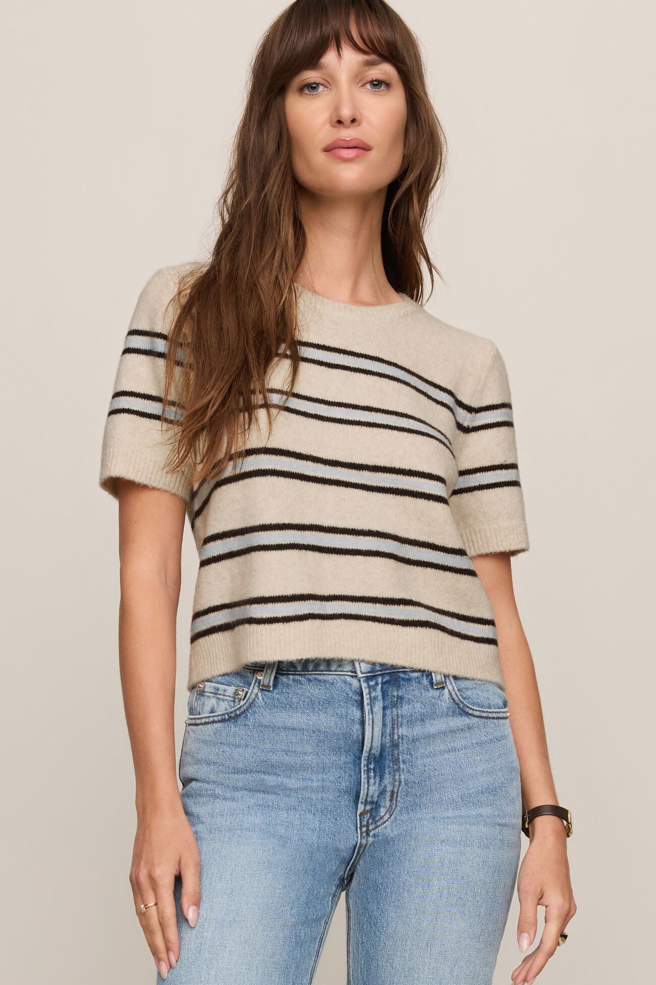 Woman wearing a beige sweater with black stripes and blue jeans on a plain background