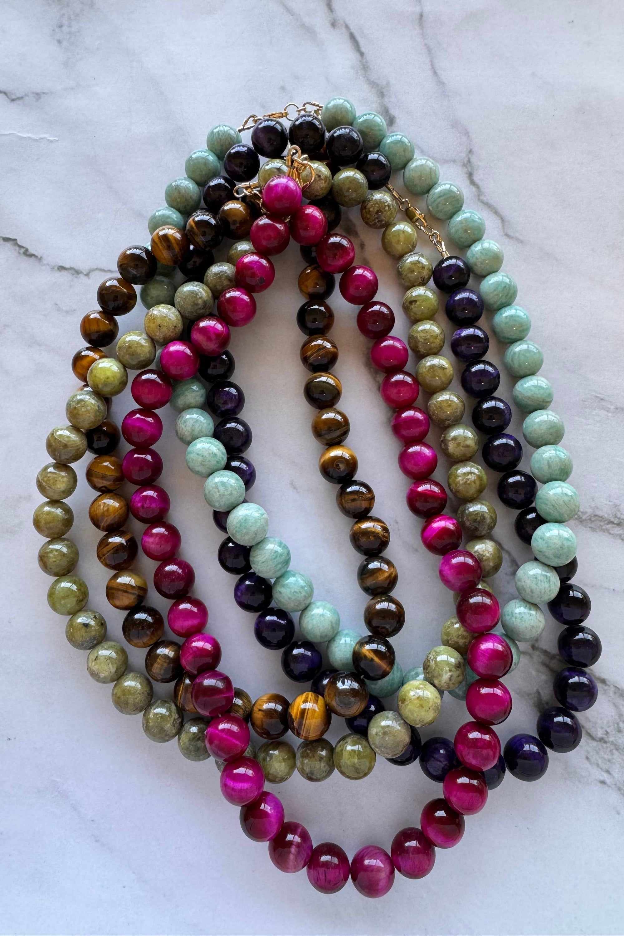 A set of multi-strand beaded necklaces arranged in a circle on a white marble surface. The necklaces feature polished round beads in vibrant magenta pink, light mint green, dark purple, brown tiger's eye, olive green, and black.