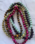 A set of multi-strand beaded necklaces arranged in a circle on a white marble surface. The necklaces feature polished round beads in vibrant magenta pink, light mint green, dark purple, brown tiger's eye, olive green, and black.