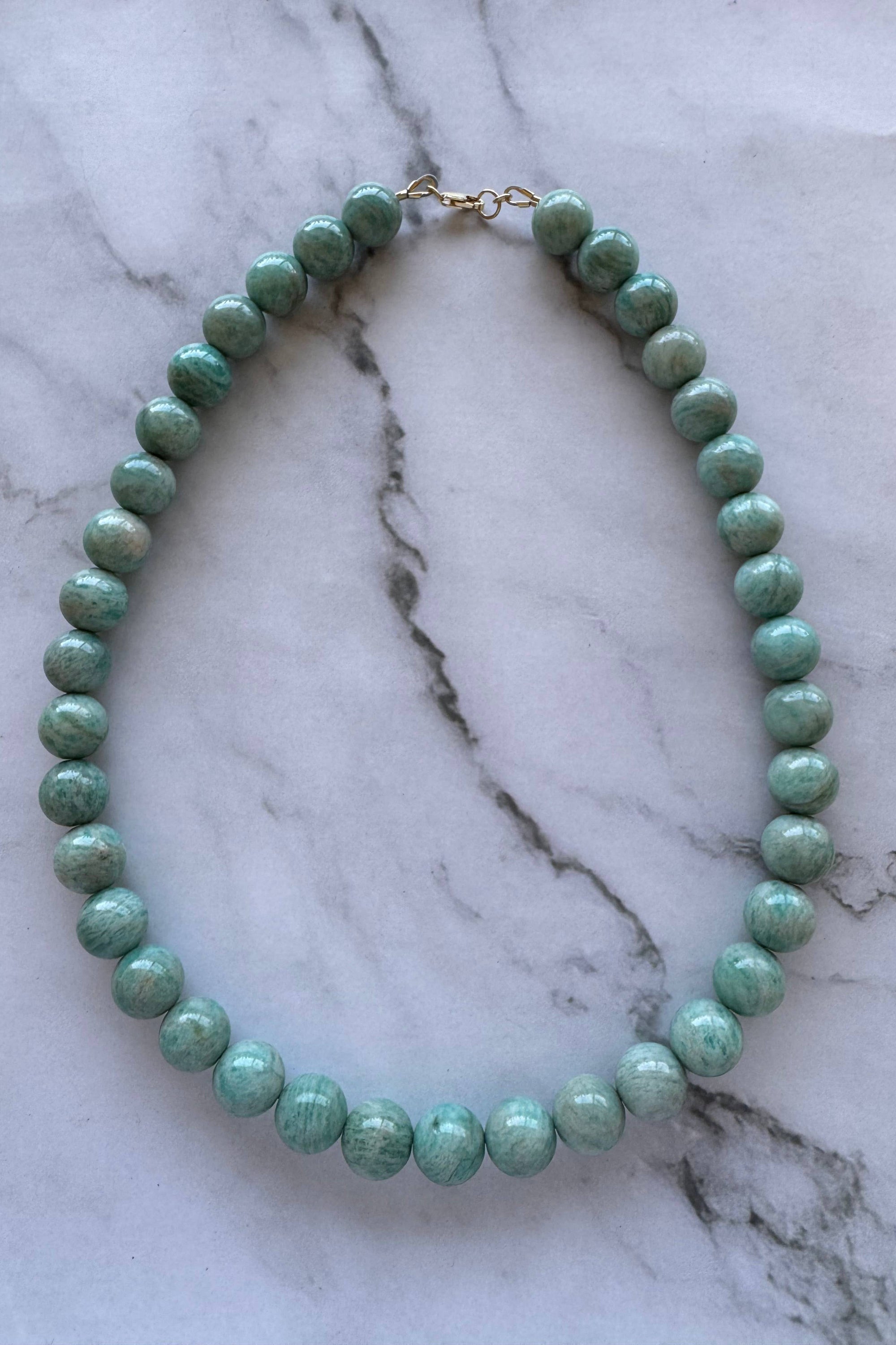 A beaded necklace made of round, polished light green beads with a small gold clasp, displayed on a white and gray veined marble surface.