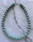 A beaded necklace made of round, polished light green beads with a small gold clasp, displayed on a white and gray veined marble surface.
