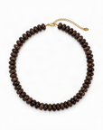 A dark brown beaded necklace made of small, polished disc-like beads, featuring a gold-tone chain extender clasp with a small tag, displayed on a clean white background.