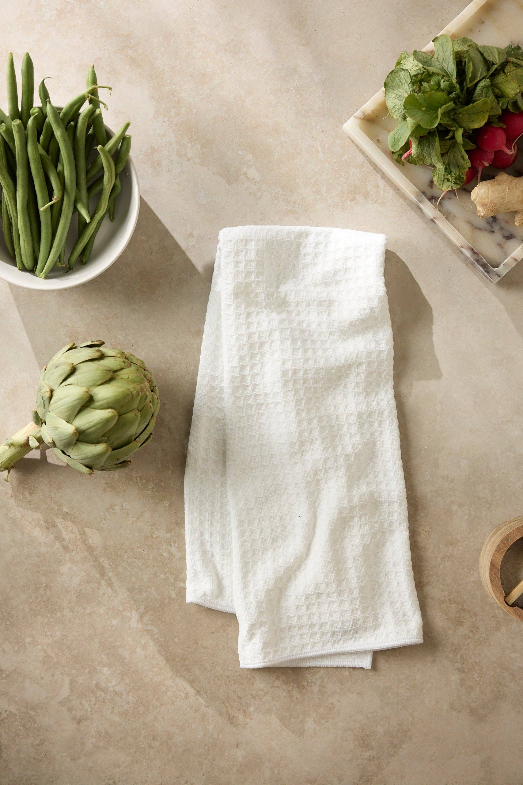 White kitchen towel on a beige surface with green beans, artichoke, and salad.